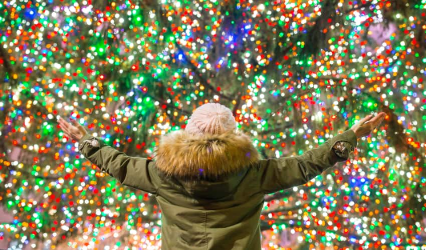 a person in a parka stands with their arms outstretched as a huge christmas tree twinkles in the background