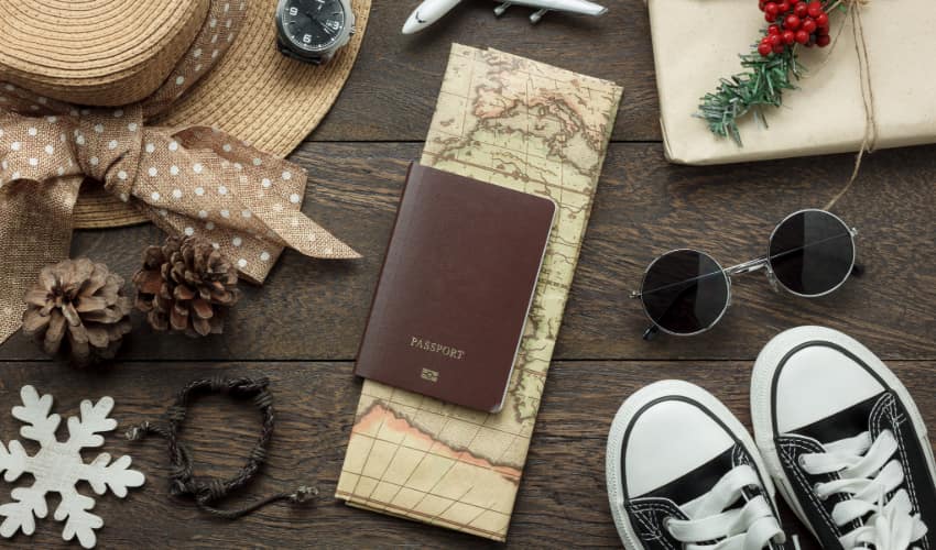 a wooden tabletop with various items spread across it: a wide-brimmed hat, a watch, a model airplane, a wrapped holiday present, sunglasses, a pair of sneakers, pine cones, a snowflake ornament, a map and a passport 