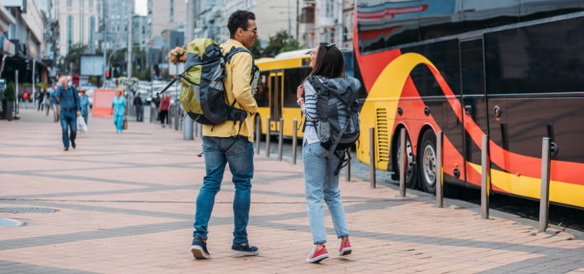 two travelers wait for their charter bus, carry-on bags in hand