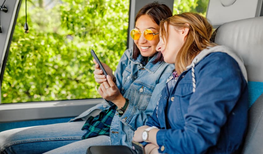 two friends relax on a charter bus, watching a video on a cell phone