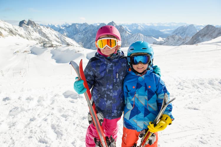 two young kids hold their skis and wear their goggles with snowy mountains in the background