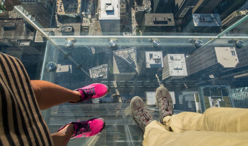 a couple stand on the Skydeck glass viewing platform in Chicago, the street visible beneath them