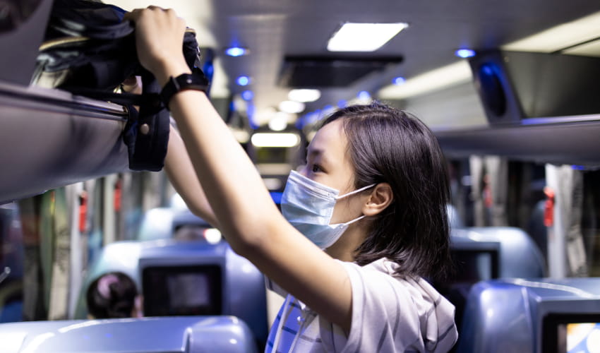 A woman wearing a mask stows her personal bag in the overhead bin of a charter bus