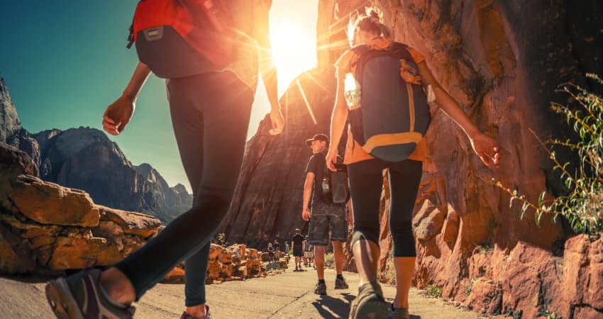 A group of hikers with backpacks walk along a paved trail in Zion National Park on a sunny day