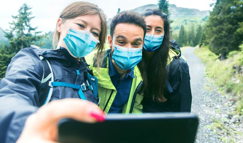 Three hikers in masks take a selfie while on the trail, snowy mountains and green trees in the background