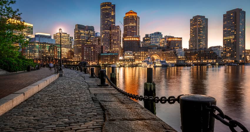The sun sets over the Boston skyline, viewed from a cobblestone walkway on the harbor