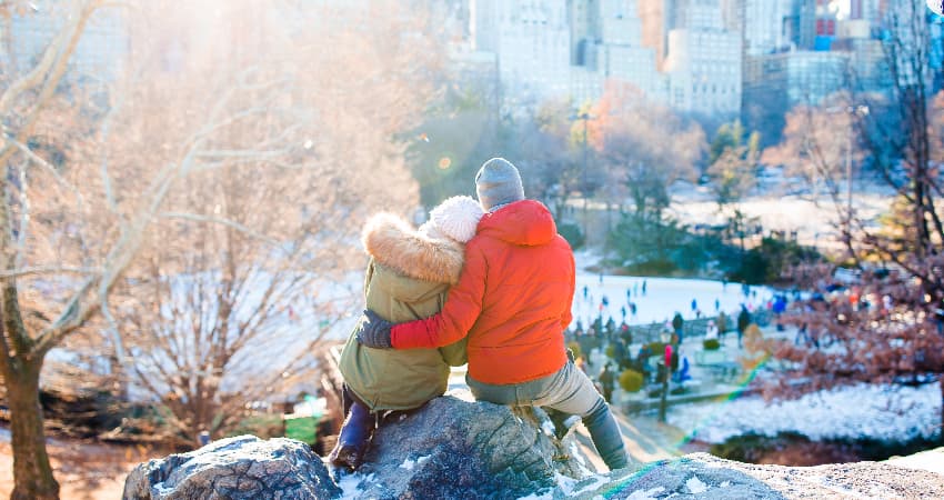 A happy couple in parkas sit on a rock outcropping in Central Park and watch ice skaters below