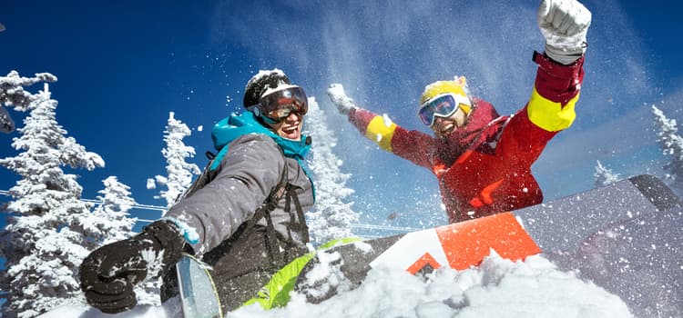 two friends hold their skies while one jumps and one smiles for the camera