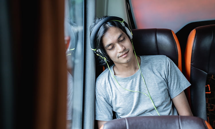 A man with headphones leans against a bus window, sleeping