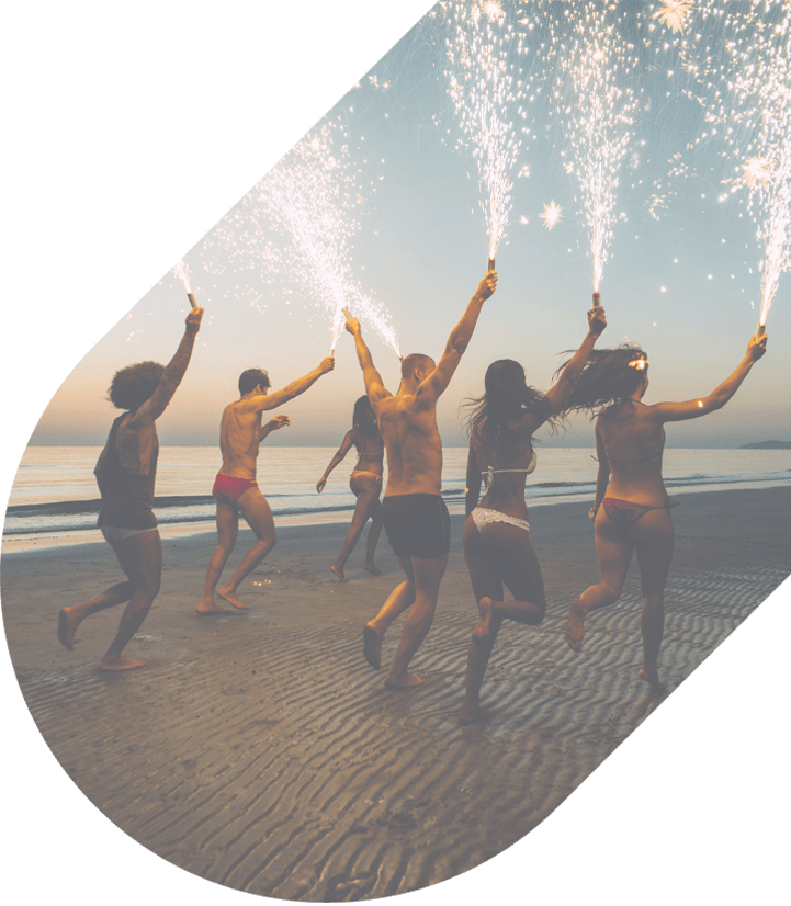 A group of party goers in bathing suits run along a beach holding sparklers