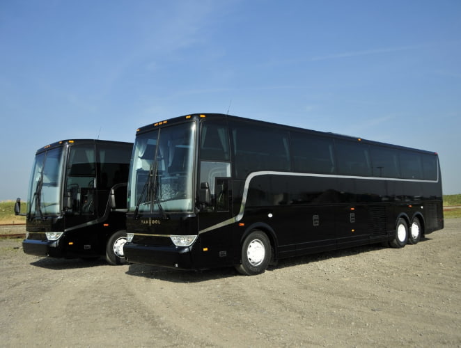 Two black Van Hool TX45 charter buses park in a dirt lot