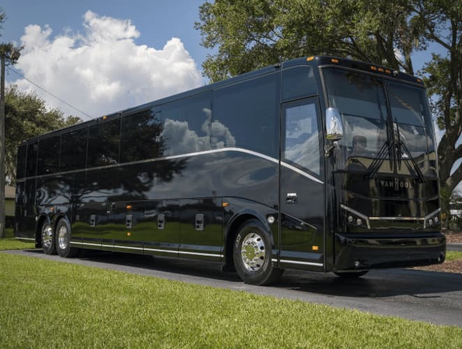 A black Van Hool CX45 charter bus parked in front of trees on a sunny day