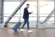 a man looks at his phone while he walks through the airport with a suitcase